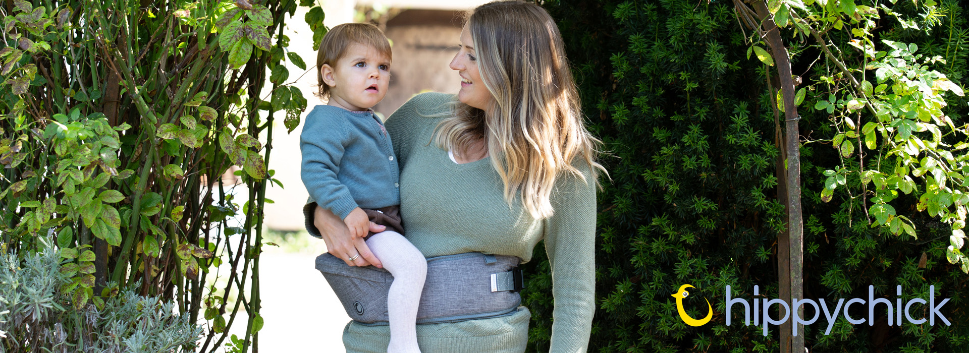 Frau trägt ein Kind im Tragegurt in einem Garten mit grünen Pflanzen.