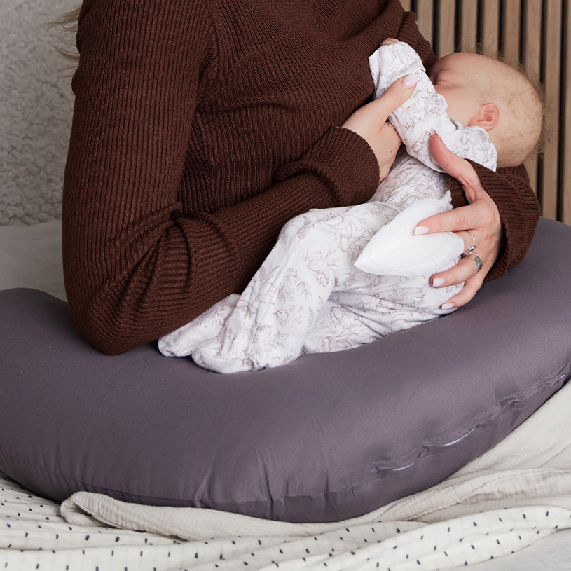Person stillt Baby mit Kissenunterstützung, brauner Pulli.