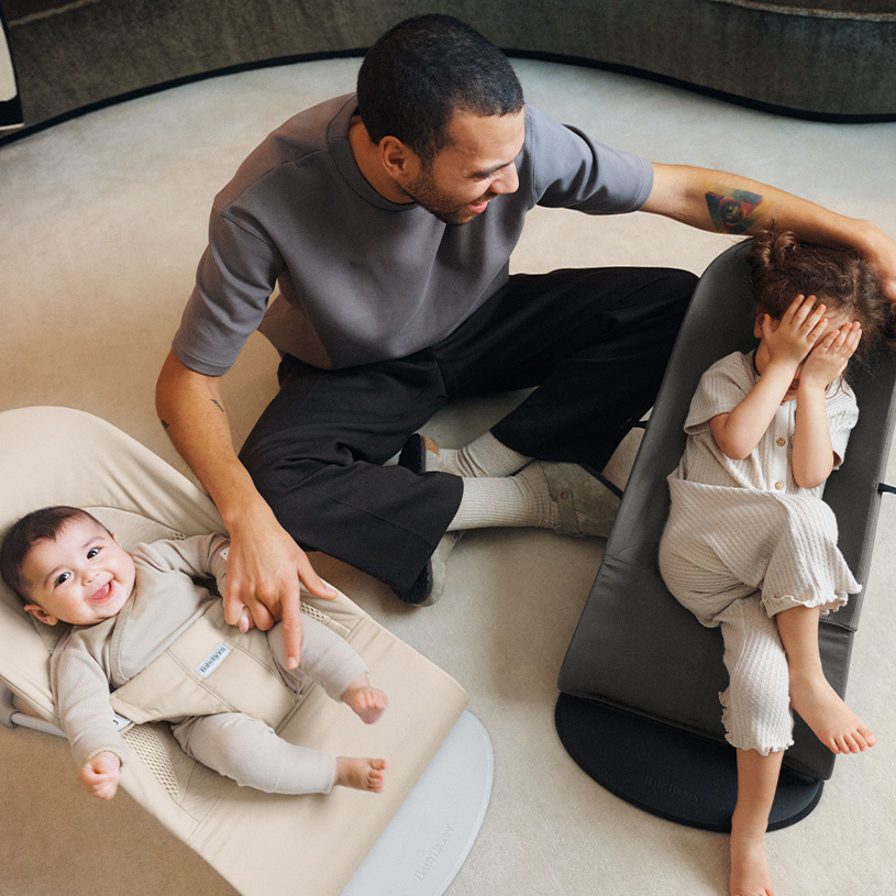 Erwachsener mit zwei Kindern, ein lachendes im Stuhl und eins spielt Verstecken.