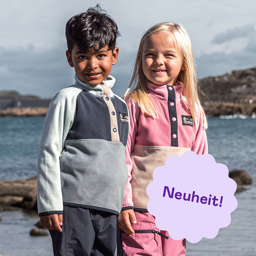 Zwei Kinder in bunten Regenmänteln auf einem Felsen am Meer, Text: Neuheit!