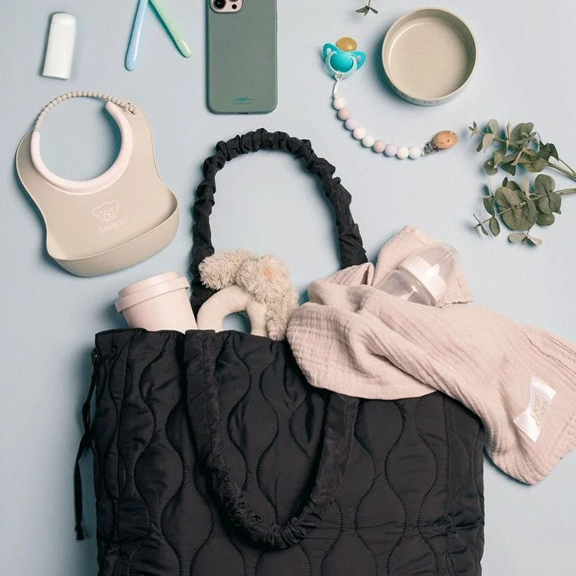 Schwarze Wickeltasche mit Babyartikeln wie Handy, Schnuller und Spielzeug auf hellem Hintergrund.
