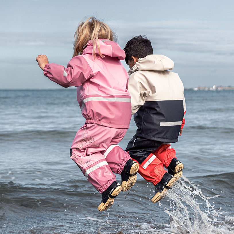 Kind in gelbem Regenhut und grüner Regenkleidung am steinigen Strand.