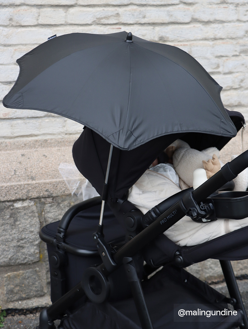 Schwarzer Kinderwagen mit Sonnenschirm, weißer Decke und Kuscheltier an Ziegelwand.