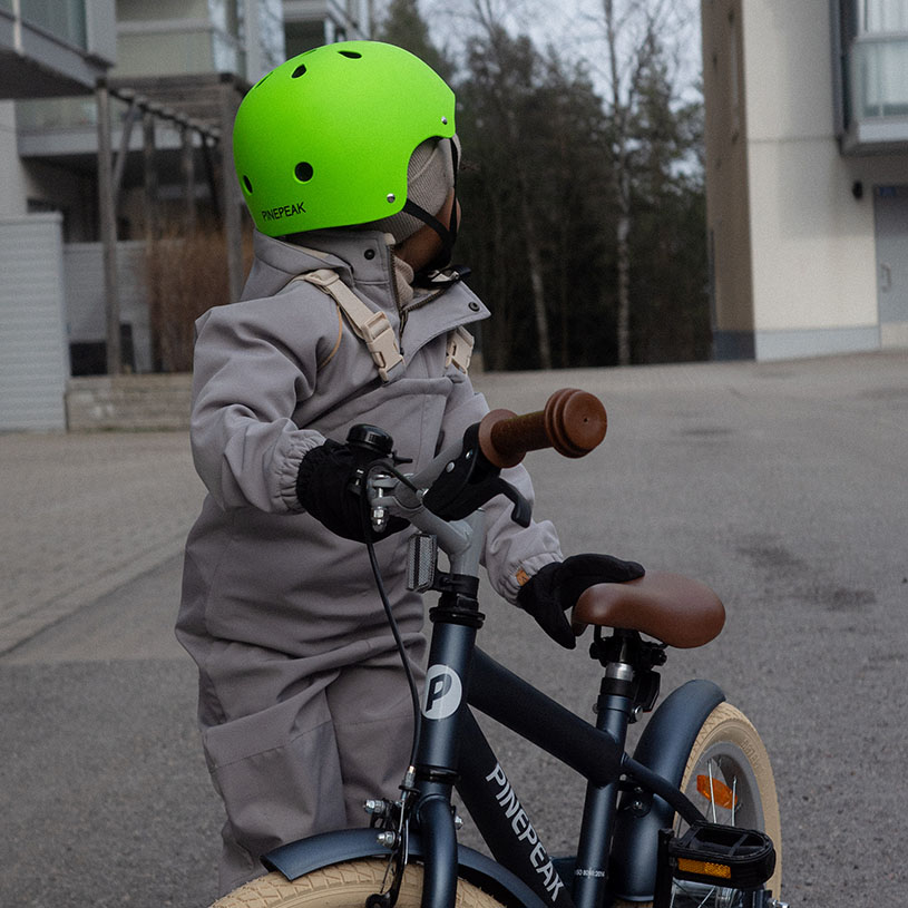 Kind auf Fahrrad, trägt grünen Helm und grauen Overall.