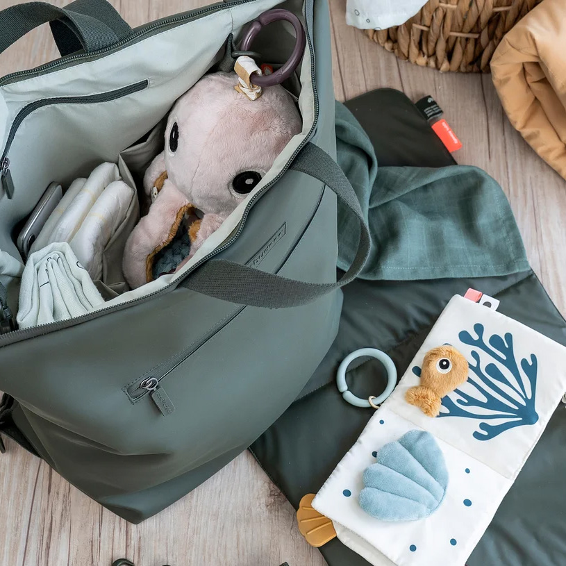 Grüne Tasche mit Babyartikeln, darunter Spielzeug und Windeln.