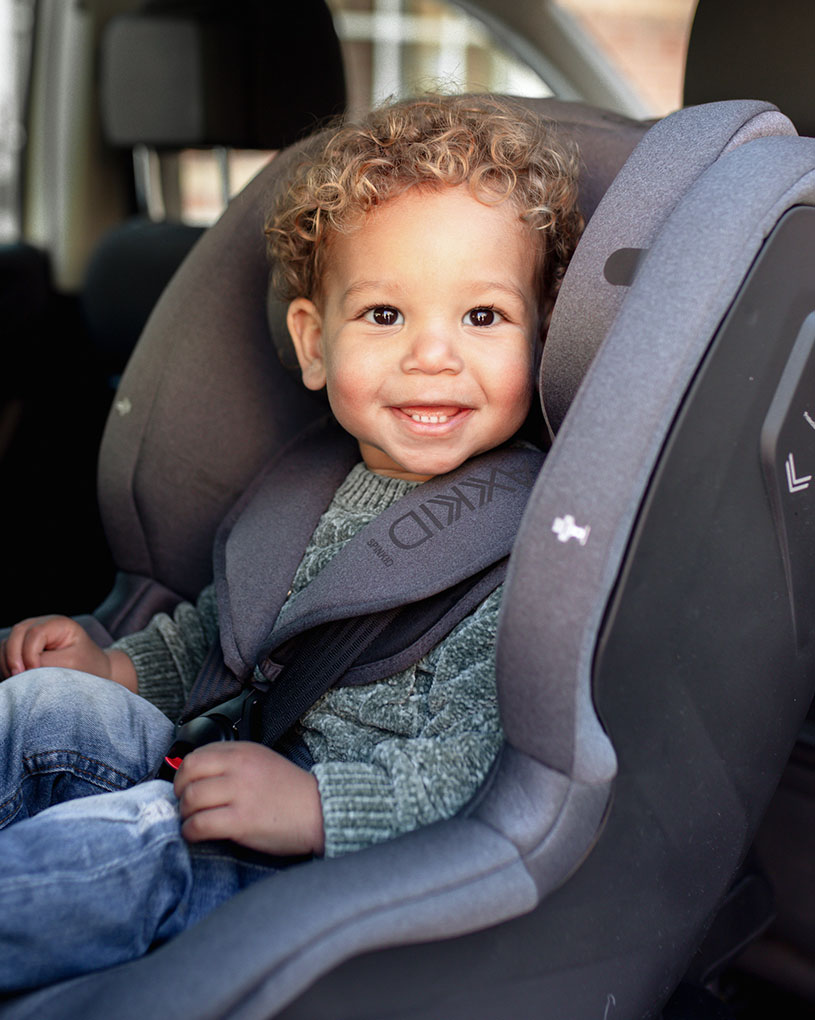 Lächelndes Kind mit lockigem Haar sitzt im Kindersitz.