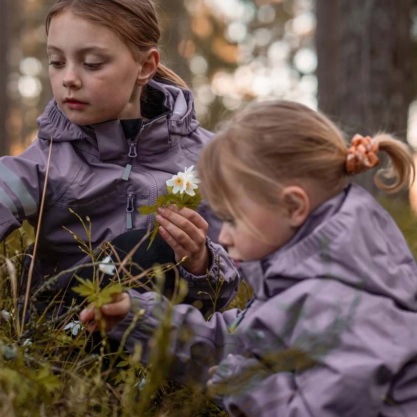 Zwei Kinder in bunten Jacken stehen lächelnd am Baum.