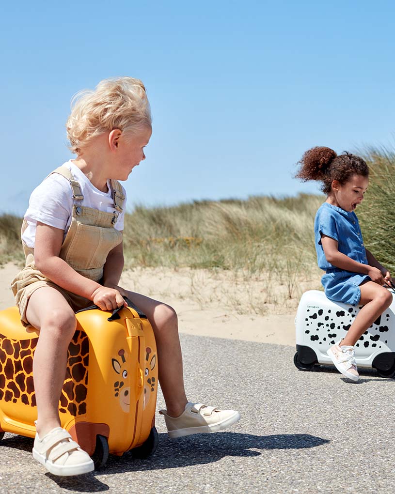 Zwei Kinder sitzen auf tiergemusterten Koffern am Strand.