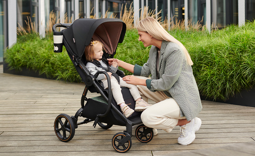 Frau lächelt Kind im Kinderwagen auf Holzterrasse mit grünen Pflanzen an.