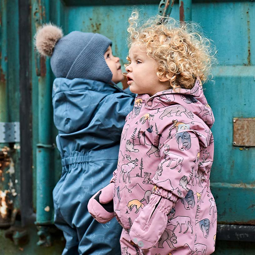 Zwei Kinder in bunten Winterkleidern stehen vor einem blauen Container.