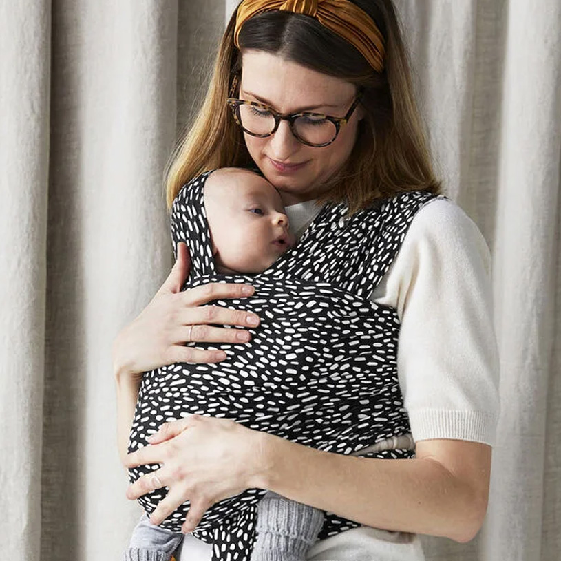Person hält ein Baby in einem schwarz-weißen Tuch vor einem Vorhang.