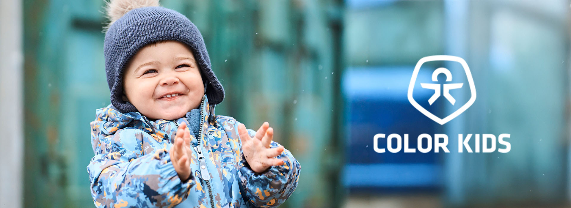 Lächelndes Kind in Winterkleidung mit blauer Mütze, Color Kids-Logo.