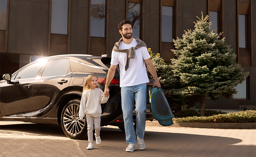Mann geht mit Kind an der Hand neben Auto, trägt Kindersitz in der Sonne.
