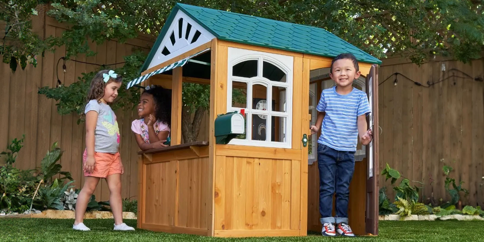 Drei Kinder spielen an einem kleinen Holzspielhaus im Garten.
