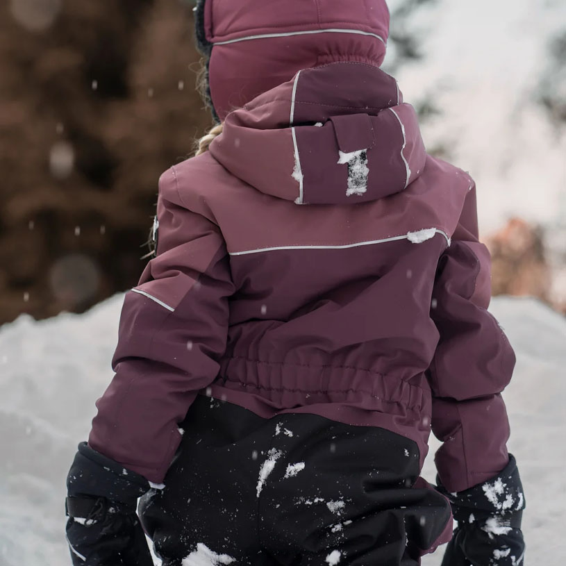 Kind in Winterkleidung sitzt im Schnee neben einem Holzzaun.
