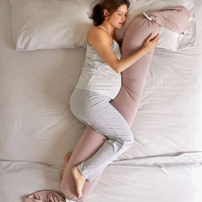 Eine schwangere Frau liegt auf einem Bett und umarmt ein langes Schwangerschaftskissen.
