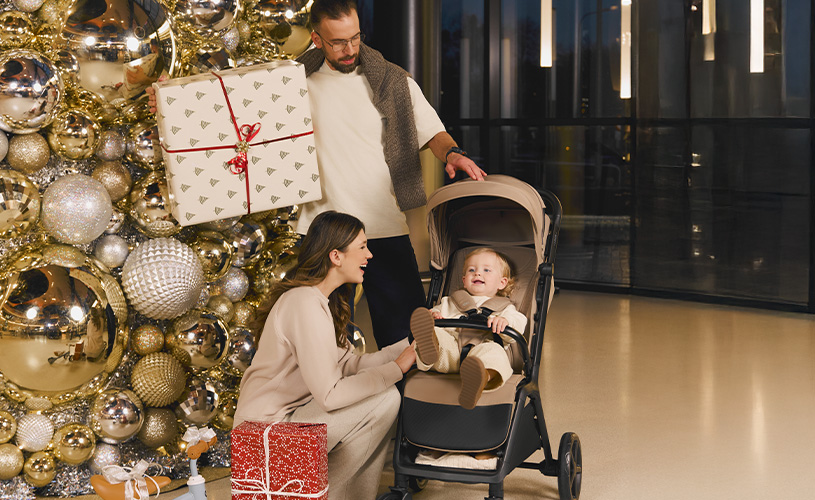 Eltern mit Kind im Kinderwagen neben Weihnachtsbaum mit Geschenken.