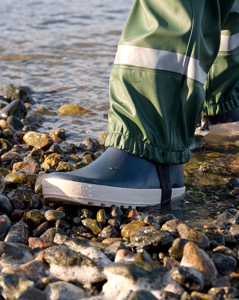 Kind in blauer Regenkleidung und rosa Schuhen geht auf Sand.