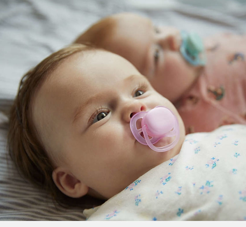 Zwei Babys liegen mit Schnullern, einer rosa, einer blau.