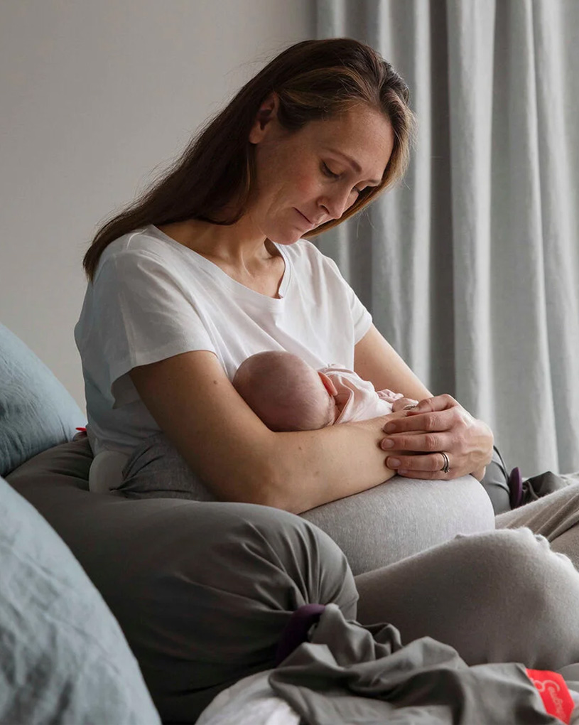 Frau in weißem T-Shirt hält ein Baby im Arm, sitzt auf grauem Sofa.
