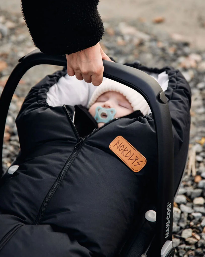 Baby in schwarzem Schlafsack in Autositz mit Schnuller, Erwachsene Hand hält den Griff.