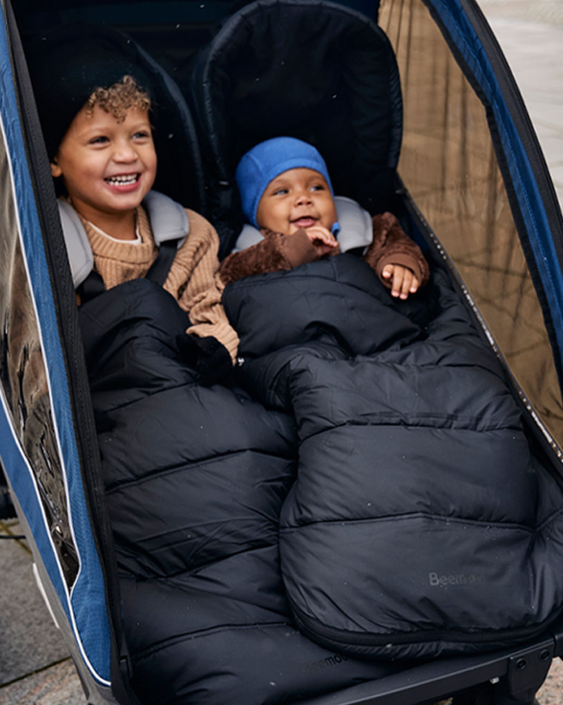 Zwei fröhliche Kinder in Schlafsäcken im Doppelkinderwagen.
